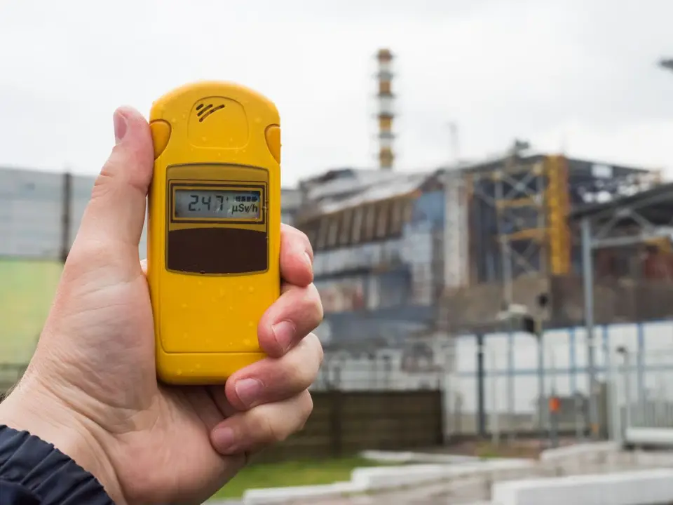 Kézben tartott sárga digitális sugárzásmérő egy ipari létesítmény előtt.